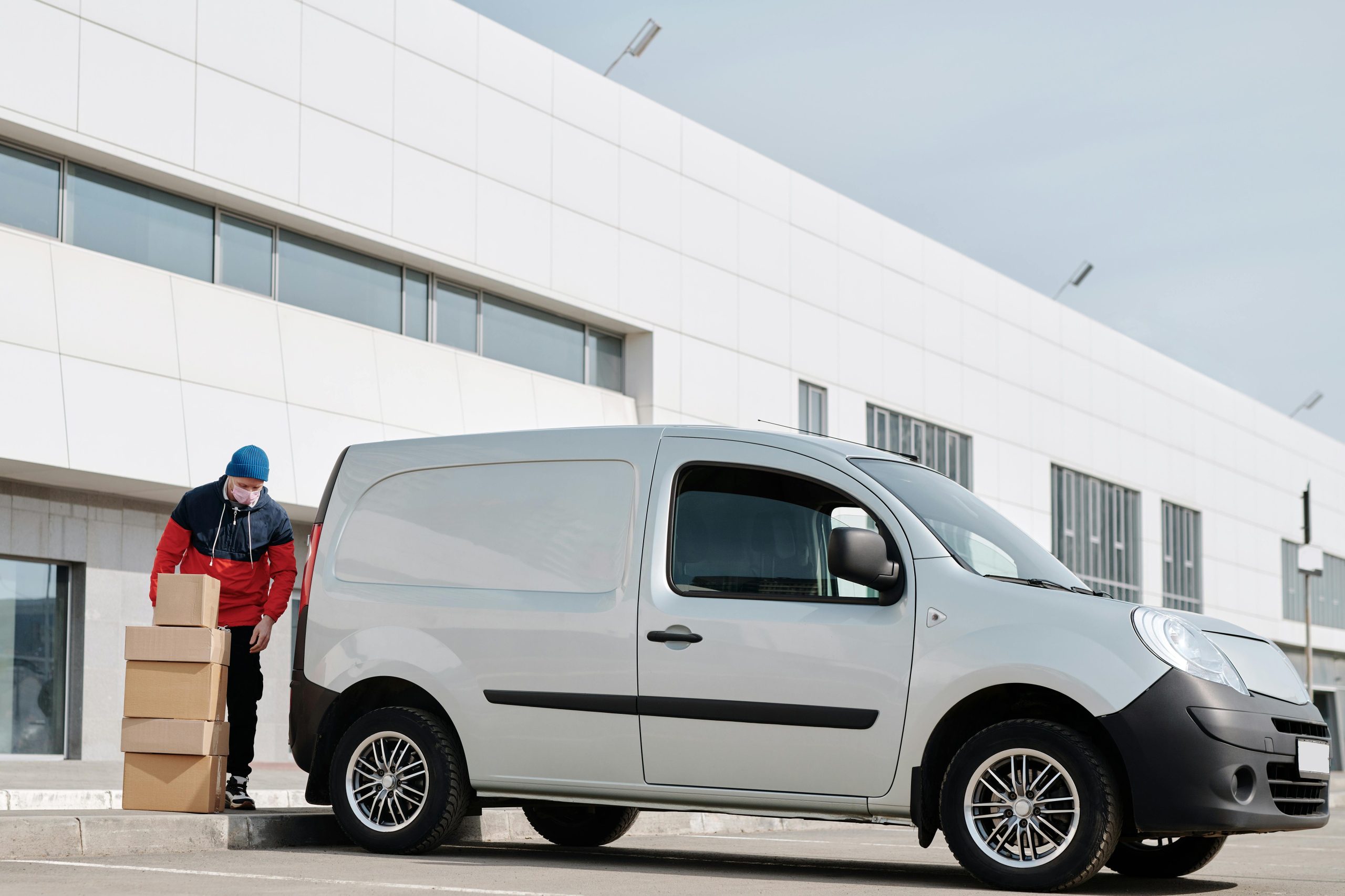 A delivery driver wearing a face mask unloads packages from a van outside a modern building.